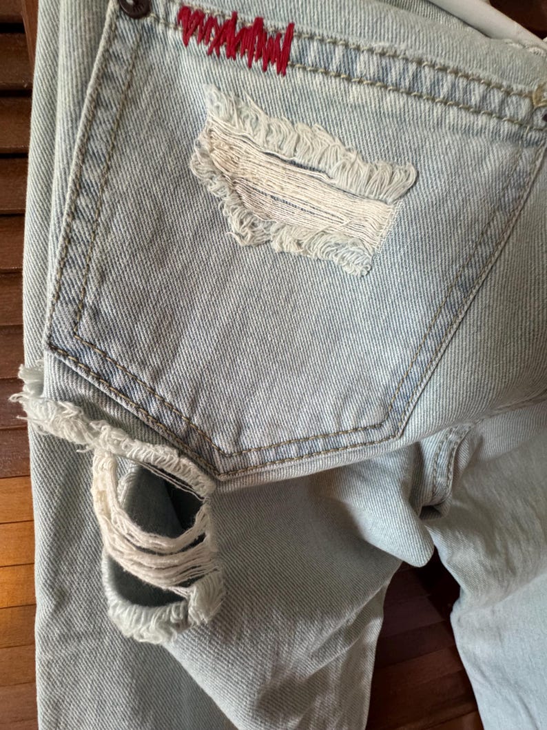 May include: Pale blue denim jeans with distressed detailing. The back pocket has a large, frayed hole and red stitching. The jeans have a classic five-pocket design and a faded wash.