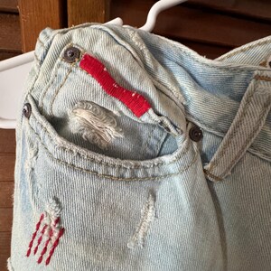 May include: Close-up of light blue denim fabric with distressed details. Features a pocket with red embroidered accents and a red fabric detail. Visible metal button closures and a white hanger.