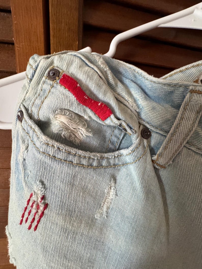 May include: Close-up of light blue denim fabric with distressed details. Features a pocket with red embroidered accents and a red fabric detail. Visible metal button closures and a white hanger.