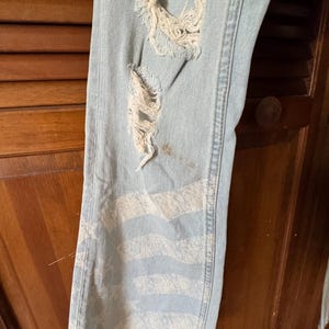 May include: Light blue denim jeans with distressed details, including ripped sections and a faded white striped design on the lower leg. The jeans have a raw, unfinished hem.