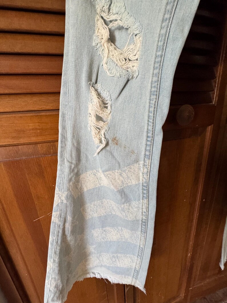 May include: Light blue denim jeans with distressed details, including ripped sections and a faded white striped design on the lower leg. The jeans have a raw, unfinished hem.
