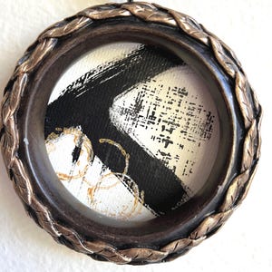 May include: A round, framed artwork with a dark brown, leaf-patterned border. The artwork inside features a black and white abstract design with gold accents. A beige ribbon is attached for hanging.