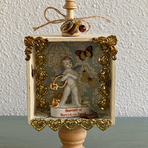 May include: A decorative box with a wooden base and a small cherub figurine. The box features gold accents, butterflies, and the text "Souvenir of Berwick-on-Tweed". A wooden chess piece is attached to the top with twine.