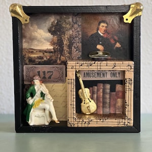 May include: A black shadow box with gold corner accents, featuring a miniature violin, a figurine of a violinist, and a painting of a violinist. The box includes the text "FOR SKILL AND AMUSEMENT ONLY".