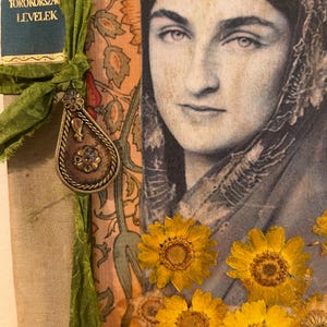 May include: A mixed media art piece featuring a black and white portrait of a person wearing a head covering, layered over a floral patterned background. Pressed yellow flowers are attached to the bottom, and a green ribbon holds a metal charm.