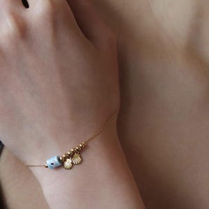 Pode incluir: Uma delicada pulseira dourada com uma conta cilíndrica azul clara, pequenas contas douradas e três pingentes de concha dourados. A pulseira é usada em um pulso, mostrando sua elegância sutil e tema praiano.