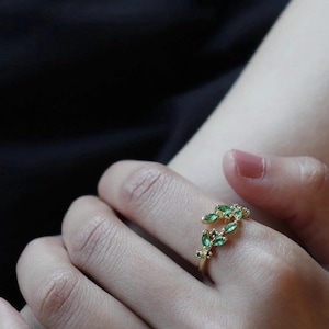 Peut inclure: Une bague dorée ornée d'un motif de feuilles avec de petites pierres précieuses vertes. La bague est portée au doigt, mettant en valeur les détails délicats du bijou. Les pierres précieuses sont d'un vert vif, ajoutant une touche de couleur.
