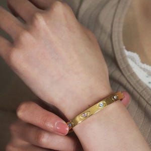 May include: Gold-colored bangle bracelet with eye-shaped designs, each containing a small, colorful gemstone. The bracelet is worn on a wrist, showcasing its polished finish and intricate details. The gemstones are pink, blue, and clear.