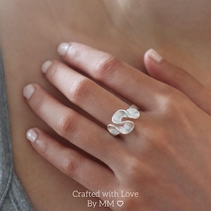 Peut inclure: Une bague en argent présentant un design fluide et continu en forme de vague qui s'enroule autour du doigt. Le métal possède une finition mate et lisse avec des bords arrondis, créant une forme sculpturale abstraite sur le doigt.