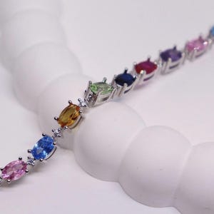 May include: A silver bracelet featuring a series of oval-cut gemstones in a rainbow of colors. The gemstones include purple, pink, blue, yellow, and green, set in a silver band. Small clear stones are interspersed between the larger gems.