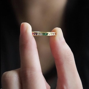 Puede incluir: Un anillo dorado con una banda de piedras preciosas de colores alternos, incluyendo rojo, morado, blanco, verde y amarillo. El anillo se sostiene entre dos dedos sobre un fondo borroso.