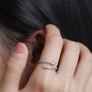 May include: A silver snake ring encircles a finger. The ring has a textured band and a snake head design with a small, round detail. The ring is worn on a hand with pink nail polish.