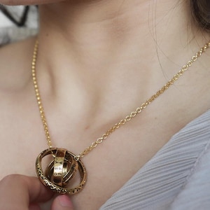 May include: A gold-colored necklace featuring a pendant with multiple interlocking rings. The pendant has engraved text and intricate detailing. The necklace is worn around the neck, with the chain visible. The overall design is ornate and elegant.