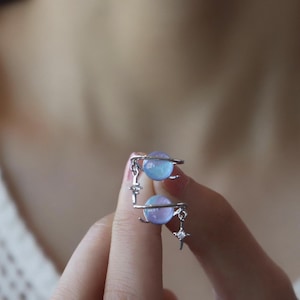 Può includere: Orecchini argentati con due sfere a forma di pianeta blu e viola. Ogni sfera è circondata da una fascia argentata, con ciondoli a forma di stella. Gli orecchini sono tenuti dalle dita di una persona.