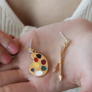 Puede incluir: Un par de pendientes dorados con temática de artista. Un pendiente es una paleta con toques de pintura roja, amarilla, negra y verde. El otro pendiente presenta dos pinceles cruzados. Los pendientes se sostienen en una mano.