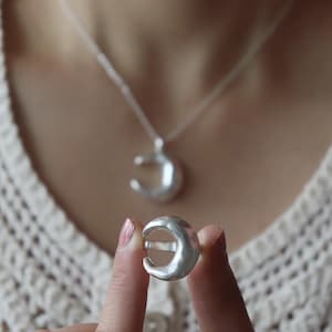 May include: A silver crescent moon ring held between fingers, with a matching crescent moon pendant necklace in the background. The ring and pendant have a textured, matte finish. The necklace hangs on a silver chain.