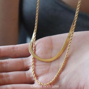 May include: A layered gold-toned necklace set consisting of two distinct chains. The top layer is a flat, flexible herringbone chain, while the bottom layer is a textured, twisted rope chain. Both chains feature a polished, metallic finish.