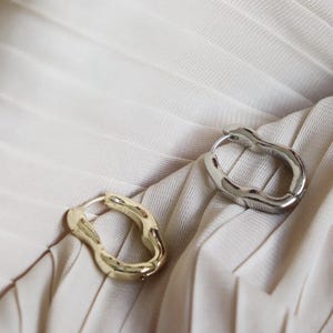 May include: A pair of hoop earrings featuring an irregular, organic, wavy shape. One earring is finished in a polished gold tone, while the other is finished in a polished silver tone. Both earrings have a smooth, reflective metallic surface.