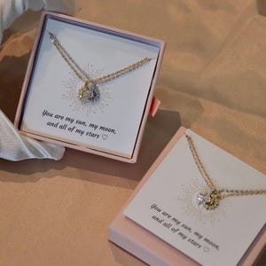Peut inclure: Un collier doré avec un pendentif soleil et lune, présenté dans un coffret rose. Le coffret contient une carte avec le texte "You are my sun, my moon, and all of my stars". Le collier est présenté sur une carte blanche.
