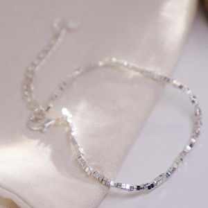 May include: A delicate silver bracelet with small, square beads. The bracelet has a simple design with a lobster clasp and an adjustable chain. The jewellery is displayed on a white surface.