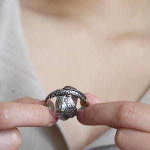 Op de afbeelding: Een zilverkleurige ring met een ingewikkeld ontwerp, vastgehouden tussen twee vingers. De ring heeft meerdere kruisende banden, met gegraveerde patronen en symbolen. De ring heeft een vintage of antieke uitstraling.