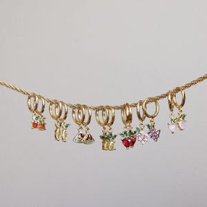 Puede incluir: Una colección de pendientes de aro dorados, cada uno adornado con abalorios de piedras preciosas de colores. Los abalorios presentan varios diseños de frutas y plantas, como cerezas, limones, sandías, fresas y plantas en macetas. Los pendientes se muestran en una cadena dorada.