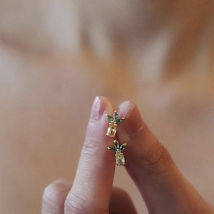 以下が含まれることがあります： ミニチュアパイナップルの形をした、金色のスタッドピアス。各イヤリングは、黄色の宝石のボディと緑色の宝石の葉が特徴です。イヤリングは、ぼやけた背景に対して指の間に保持されています。