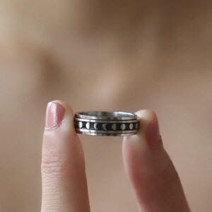 May include: A silver-colored ring with a black band featuring a series of moon phase designs. The ring is held between two fingers against a blurred background.