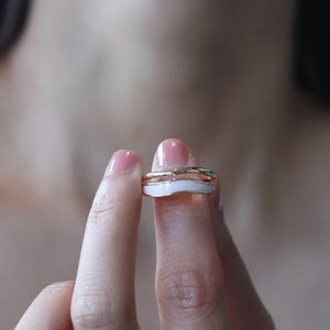 Könnte beinhalten: Zwei goldene Ringe mit einem weißen Emaille-Wellenmuster. Die Ringe sind übereinander gestapelt und zeigen eine minimalistische und moderne Ästhetik. Die Ringe werden von den Fingern einer Person gehalten.