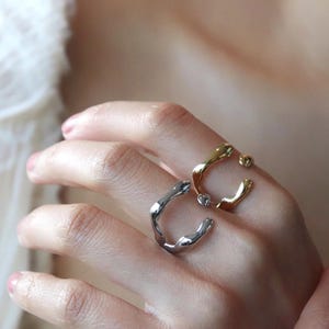 Può includere: Tre anelli astratti nei toni dell'oro e dell'argento sono esposti su una mano. Gli anelli hanno un design organico e fluido. Un anello ha una piccola gemma trasparente. Gli anelli sono un gioiello unico.