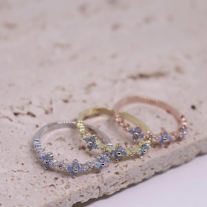 May include: Three delicate rings in silver, gold, and rose gold, each adorned with small, light blue floral designs. The rings are displayed on a textured, light-colored surface, showcasing their intricate details.