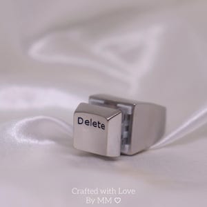 Puede incluir: Un anillo plateado con diseño de tecla de ordenador con la palabra "Delete" impresa. El anillo tiene forma rectangular y acabado pulido. El anillo está sobre un fondo blanco suave.