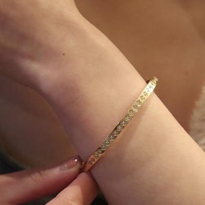 May include: A gold-coloured bangle bracelet featuring a repeating pattern of small, clear gemstones. The bracelet is worn on a person's wrist. The design is visible. The bracelet appears to be made of metal and is a jewelry item.