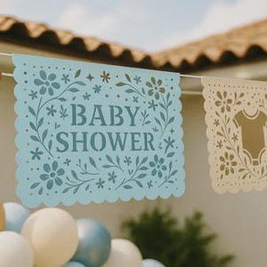以下が含まれることがあります： スカラップエッジのライトブルーの紙製バナー。ダークブルーのフォントで「BABY SHOWER」という文字が記され、複雑な花の切り抜きが施されています。バナーは白い紐で吊るされており、花やベビー服の柄が描かれた他の装飾パネルと並んでいます。