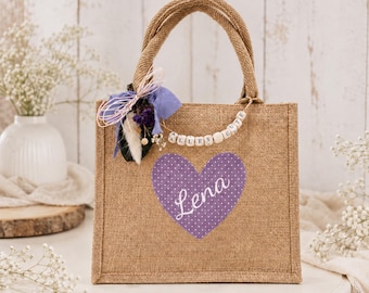 May include: A tan jute tote bag featuring a purple heart with white polka dots and the name Lena printed in white script. The bag is decorated with a purple ribbon, dried flowers, and a string of wooden beads spelling out ALLES GUTE.