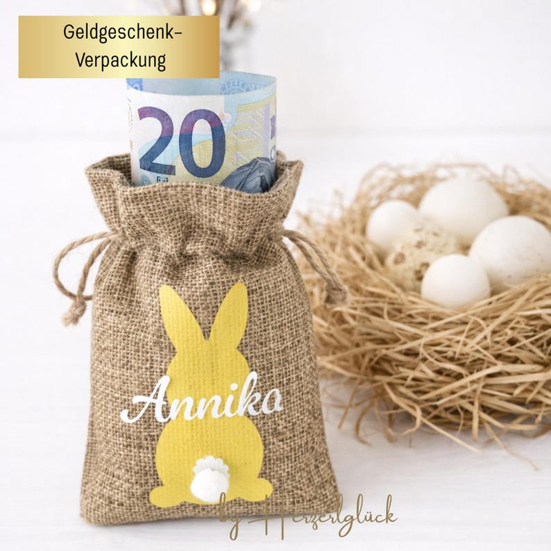 May include: A burlap gift bag with a yellow bunny design and the name "Annika" in white. The bag contains a rolled-up twenty-dollar bill. A nest with eggs is in the background. The text "Geldgeschenk-Verpackung" is at the top.