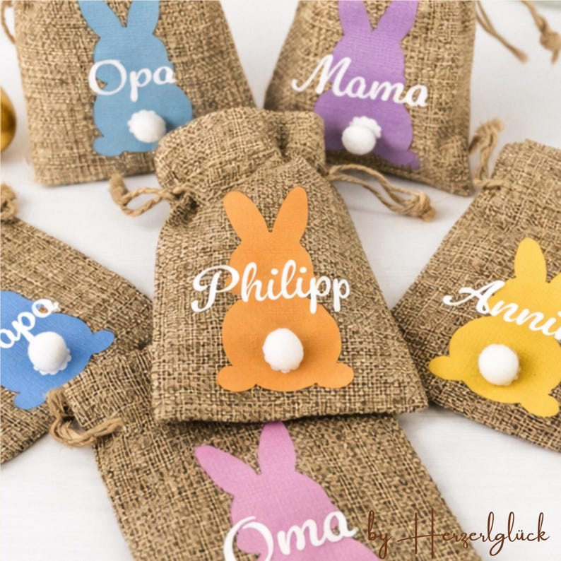 May include: Burlap drawstring bags with colorful bunny cutouts and white pom-pom tails. Each bag features a name in white script, perfect for Easter gifts or party favors. The bunny cutouts are in blue, purple, orange, and yellow.