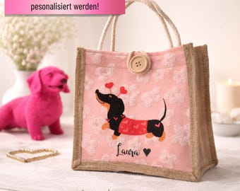 May include: Pink tote bag with a dachshund illustration wearing a red sweater with heart accents. The bag has a floral pattern and a button closure. The text "Laura" and a heart are below the dachshund. The bag has jute trim and handles.