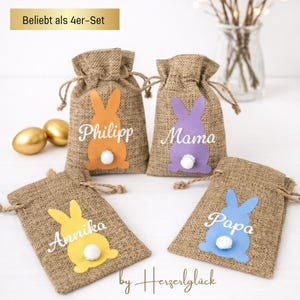 May include: Four burlap Easter gift bags with bunny silhouettes in orange, purple, yellow, and blue. Each bag has a white pom-pom tail and a name: Philipp, Mama, Annika, and Papa. The text "Beliebt als 4er-Set" is at the top.