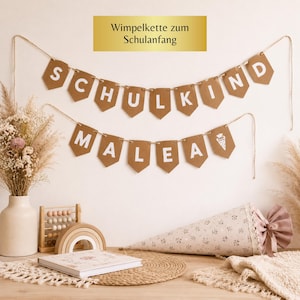 Könnte beinhalten: Dekorative Wimpelkette mit den Wörtern "SCHULKIND" und "MALEA" in weißen Buchstaben auf braunen Wimpeln. Die Kette hängt an einer weißen Wand. Eine Schultüte und andere dekorative Gegenstände befinden sich auf einer Holzoberfläche.