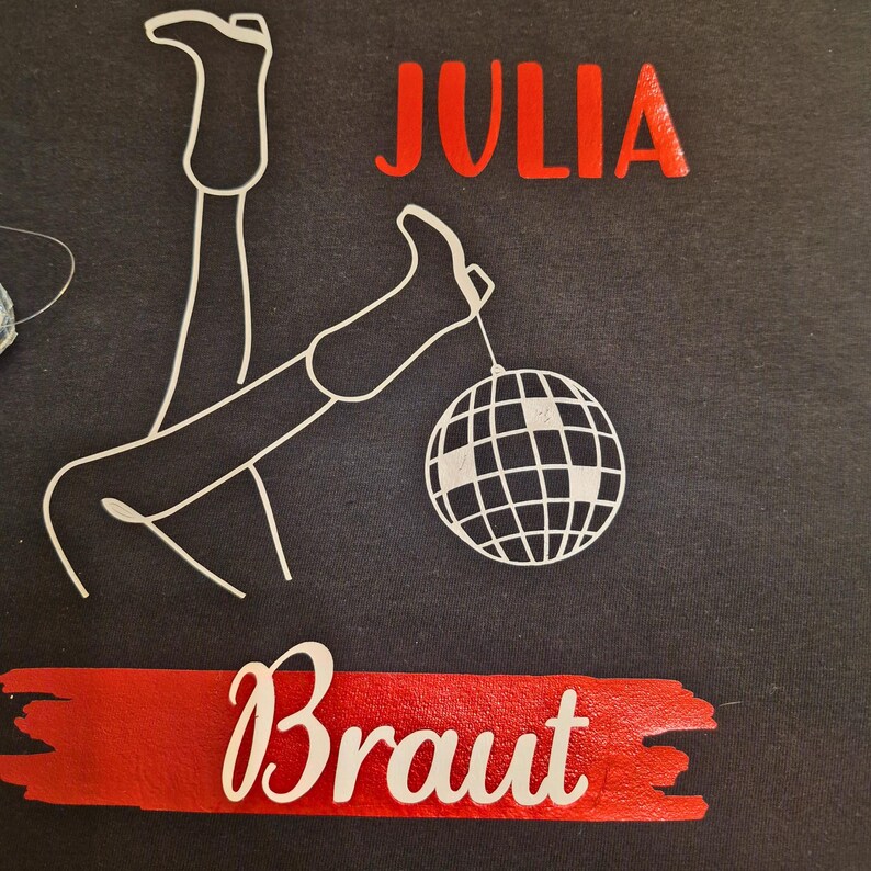 May include: Black t-shirt with white line art of legs and a disco ball. The word "JULIA" is in red above the legs, and the word "Braut" is in white on a red banner below.