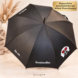 Könnte beinhalten: Ein schwarzer Regenschirm mit dem weißen Text MARLA, So ein ... und Hundewetter. Er zeigt eine Grafik eines weißen Hundegesichts mit roten herzförmigen Sonnenbrillen. Ein goldenes Etikett oben trägt die Aufschrift Regenschirm personalisierbar mit deinem Wunschnamen.