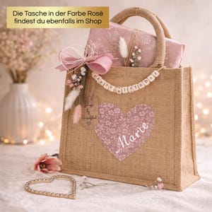 May include: A burlap tote bag with a floral heart design and the name "Marie". The bag is decorated with a pink bow, dried flowers, and a wooden bead banner that says "HAPPY BIRTHDAY". A gift is inside the bag.