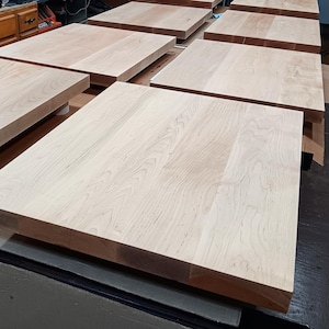 May include: Several unfinished, light-colored wooden table tops are arranged on a surface. The wood grain is visible, and the tops appear to be square or rectangular. The tables are ready for finishing.