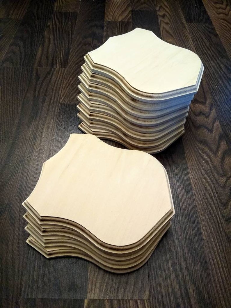 May include: A stack of unfinished wooden plaques shaped like shields. The plaques are light brown and have a smooth surface.
