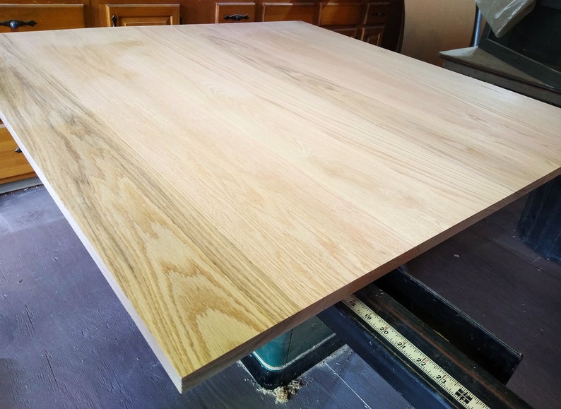 May include: A large, light-colored wood tabletop with a natural wood grain pattern. The tabletop is resting on a black and green woodworking tool.