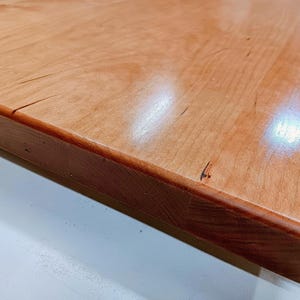 May include: Close-up of a wooden tabletop with a reddish-brown finish. The wood grain is visible, and the edges are rounded. The surface has a glossy sheen, reflecting light. The corner of the table is visible.