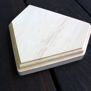 May include: A plain, unfinished wooden home plate, with a slightly rounded edge, sits on a dark wood surface.