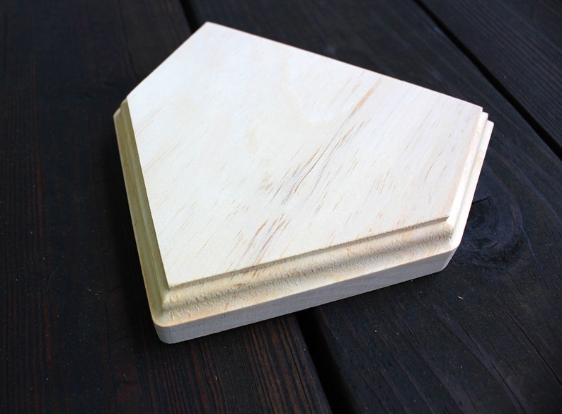 May include: A plain, unfinished wooden home plate, with a slightly rounded edge, sits on a dark wood surface.