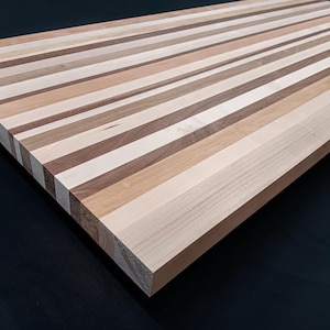 May include: A rectangular wooden cutting board featuring a striped pattern composed of alternating light, medium, and dark brown wood planks. The surface displays a smooth, flat finish with visible natural wood grain textures running lengthwise.
